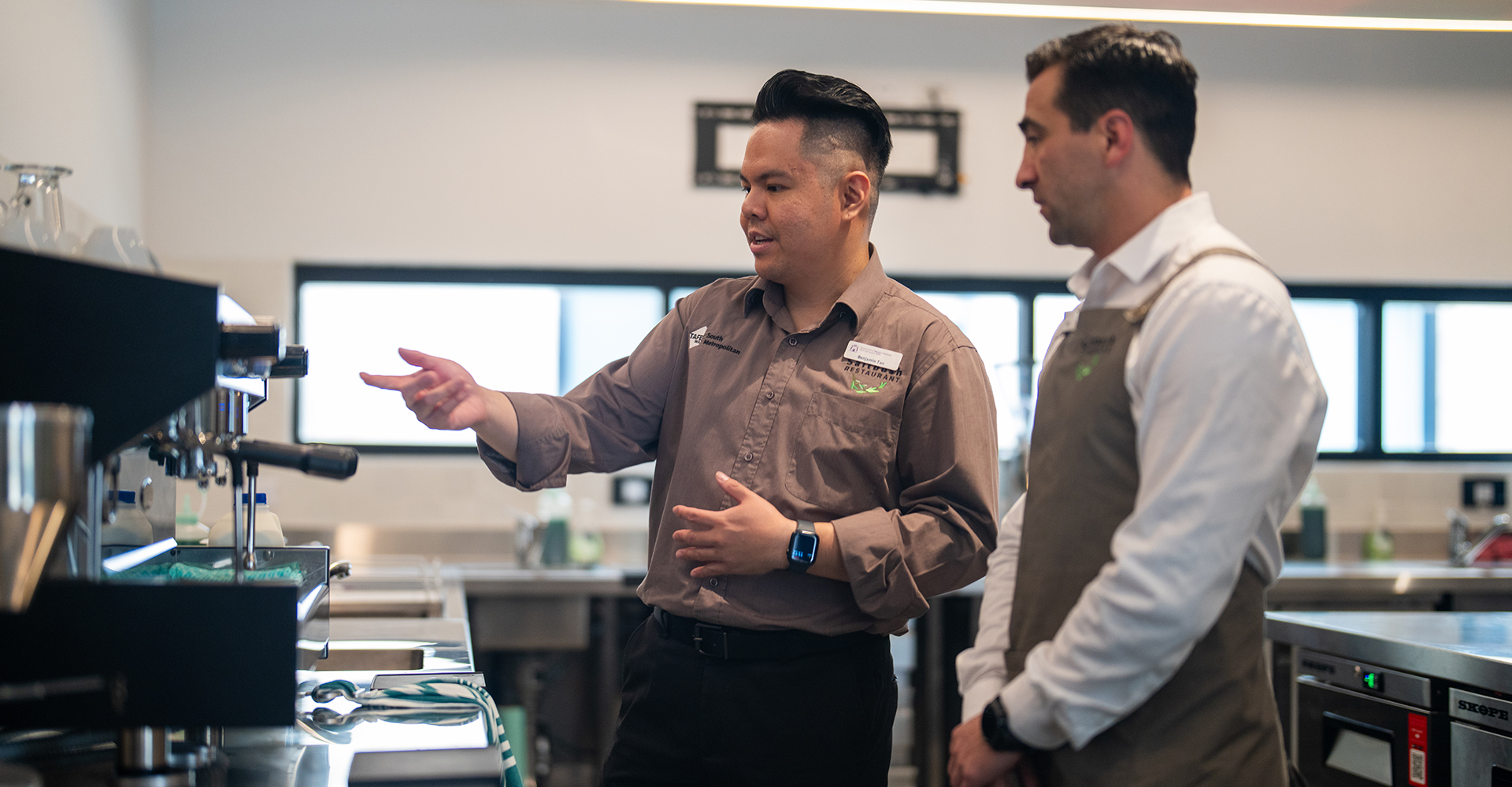 Lecturer Ben Tan point towards a coffee machine to his right. On his left, is a student wearing a white shirt and grey/brown apron.