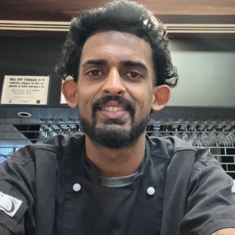 Mauritian student Dowshan is taking a selfie, looking towards the camera smiling. He has black hair, a black beard and brown eyes. He wears a black chef uniform. In the background is a closed cafeteria.