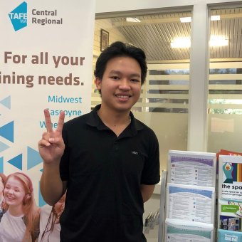 Vietnamese student Duy Ngoc Ha stands in the middle of the photo, facing towards the camera. He has black hair, brown eyes and wears a black polo shirt. He is smiling. With his right hand he is giving the peace sign. In the background of the image is a banner promoting Central Regional TAFE. On the banner it says "For all your training needs." On the student's left is a display of different flyers. Behind the student is an office.