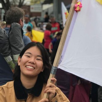 Japanese Female Student faces towards the camera with a big smile. In her hands is a Japanese flag that has been decorated with origami birds - red, yellow and blue. In the background is a crowd of people. This photo was taken at the International Festival in Bunbury.