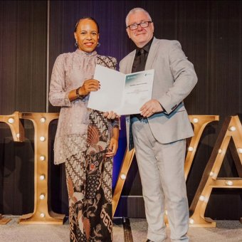 Student Herlina Yawang and North Metro TAFE's Darren Channell stand on stage at graduation, looking towards the camera. They are holding Herlina's graduation certificate. In the background there is a "TIWA" in lights and balloons on the stage.