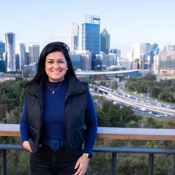 Student stands towards the camera smiling, in the background is a scenic view of the Perth city skyline.