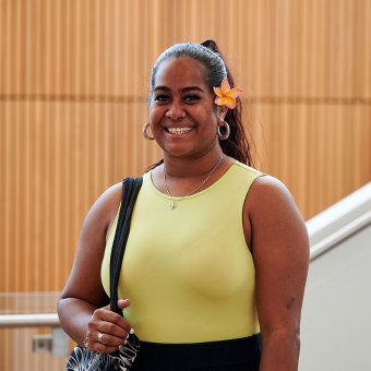 Student Keloha stands facing the camera with a big smile. She wears a yellow top and a yellow frangipani is tucked behind her ear. Behind her is a white staircase and a wall with wooden pannels.