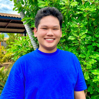 Vietnamese student Nguyen Minh Dao faces towards the camera with a big smile. He stands in front of lush vibrant greenery. He wears a bright blue t-shirt.