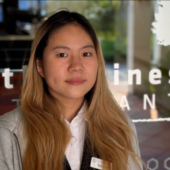 Student Nachi stands out the front of a glass window. She has blonde hair, wears a white shirt and her name badge is clipped onto her shirt. She faces the camera with a smile.