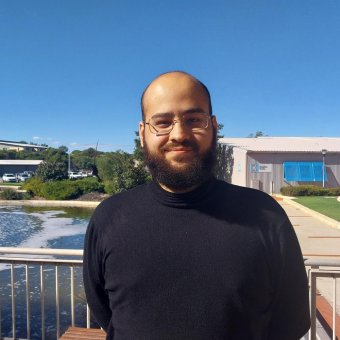 Brazilian student Pedro Vicente de Carvalho stands slightly to the right of the photograph smiling towards the camera. He has a dark brown beard, brown eyes, he wears glasses and a black long sleeve turtle neck shirt. In the background is a water fountain that sits among green bushes. Also in the background is a building made out of stainless steel sheets, it has bright blue window covers. Further in the distance is a carpark circled by tall trees.