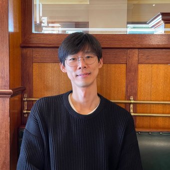 Image of smiling student Park Pilsu. He is faced towards the camera. He wears a black jumper. Behind him is a wooden wall and a mirror.