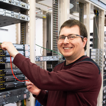 Student stands in front of information technology hardware. He smiles towards the camera.