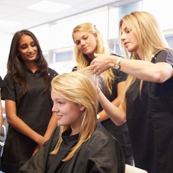 Three people stand watching a hairdresser demonstrate as they cut client's hair