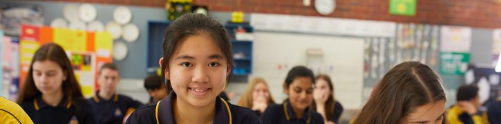 Student smiling in secondary schools classroom, with other children seated at desks in background