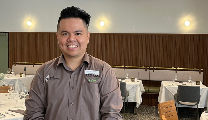 Ben Tan faces the camera smiling. He has brown hair and brown eyes and wears a formal grey button shirt. In the background is a restaurant. There's tables with white table cloths draped over the top. Glassware is placed on the tables fashionably. There are chairs tucked into the tables neatly. On the back wall, the bottom half if covered with wood whilst the top half is a white wall.