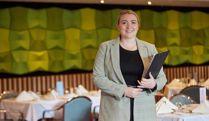Student Camille facing the camera holding a clipboard. She stands in an empty restaurant with the tables set for service, with a white tablecloth and cutlery. Behind her is a green wall feature and wooden pannels.