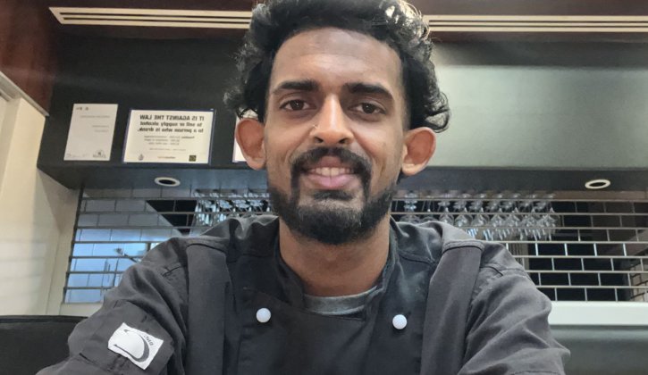 Mauritian student Dowshan is taking a selfie, looking towards the camera smiling. He has black hair, a black beard and brown eyes. He wears a black chef uniform. In the background is a closed cafeteria.