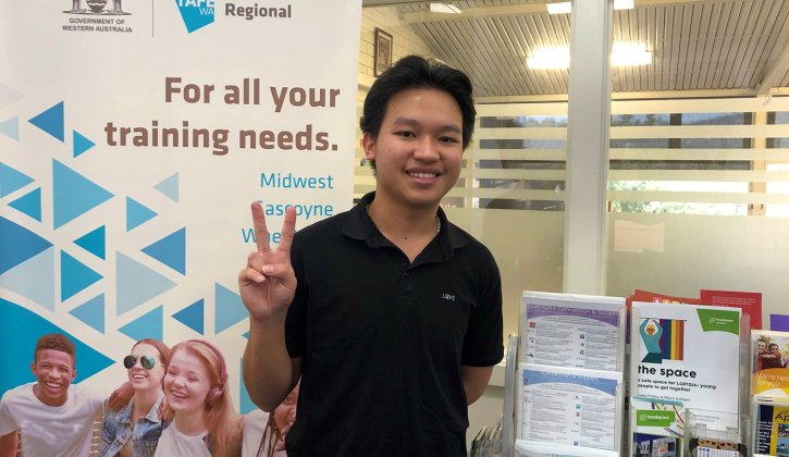Vietnamese student Duy Ngoc Ha stands in the middle of the photo, facing towards the camera. He has black hair, brown eyes and wears a black polo shirt. He is smiling. With his right hand he is giving the peace sign. In the background of the image is a banner promoting Central Regional TAFE. On the banner it says "For all your training needs." On the student's left is a display of different flyers. Behind the student is an office.