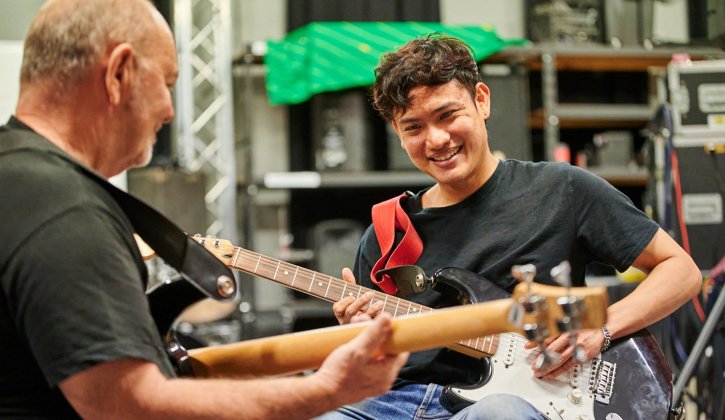 Student Farook (George) is seated holding an electric guitar. He faces a man who also holds an electric guitar. Farook is smiling at the instructor as he strums.