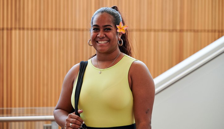 Student Keloha stands facing the camera with a big smile. She wears a yellow top and a yellow frangipani is tucked behind her ear. Behind her is a white staircase and a wall with wooden pannels.