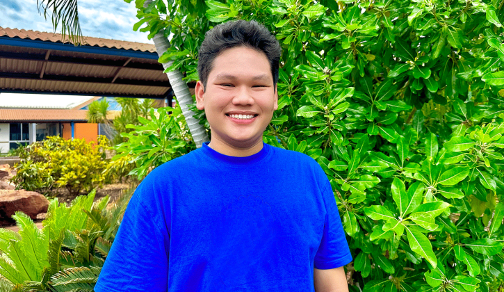 Vietnamese student Nguyen Minh Dao faces towards the camera with a big smile. He stands in front of lush vibrant greenery. He wears a bright blue t-shirt.