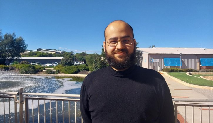 Brazilian student Pedro Vicente de Carvalho stands slightly to the right of the photograph smiling towards the camera. He has a dark brown beard, brown eyes, he wears glasses and a black long sleeve turtle neck shirt. In the background is a water fountain that sits among green bushes. Also in the background is a building made out of stainless steel sheets, it has bright blue window covers. Further in the distance is a carpark circled by tall trees.