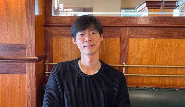Image of smiling student Park Pilsu. He is faced towards the camera. He wears a black jumper. Behind him is a wooden wall and a mirror.