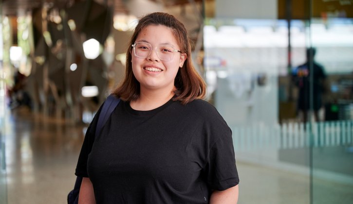 Student Athena standing in foyer of Perth TAFE campus. She wears a black t-shirt and a backpack on one shoulder. She faces towards the camera with a big smile.