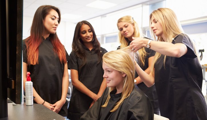 Three people stand watching a hairdresser demonstrate as they cut client's hair