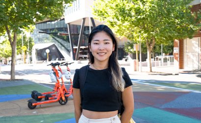 Zaki is the centre of the image. She wears shorts and a t-shirt. She smiles towards the camera. In the background is a few electric scooters, lots of big green trees and North Metropolitan campus. The pavement is colourful with red, blue and yellow squares.