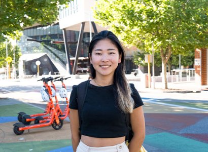 Zaki is the centre of the image. She wears shorts and a t-shirt. She smiles towards the camera. In the background is a few electric scooters, lots of big green trees and North Metropolitan campus. The pavement is colourful with red, blue and yellow squares.