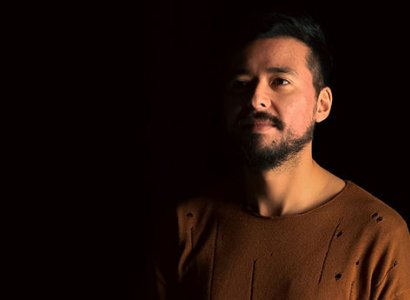 Student Pedro is on the right of the photo. He's positioned on an angle and he smiles to the distance. The lighting is dim and moody. A soft yellow light comes from the right of the image, lighting up the side of Pedro. He wears a brown shirt, he has black hair hair, brown eyes and a black beard.