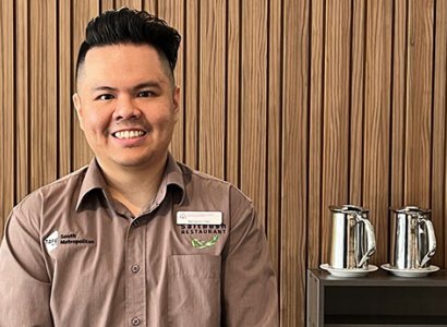 Lecturer Ben Tan stands towards the camera with a big smile. He's wearing a light brown business shirt with his name badge on his right chest above branding that says &quot;Saltbush Restaurant.&quot; On the left side of his shirt, it reads &quot;South Metropolitan&quot; with the &quot;TAFE WA&quot; logo. Sitting on a bench behind him are two plunger coffee pots in stainless steel. There's a brown panelled wall that fills the backdrop.