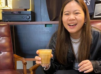 Nachi is situated to the right of the image, she's laughing and looking towards the camera. In her right hand is a latte coffee. She's sitting on a brown leather seat with another brown leather seat on her right that's empty. There's a bench in the background with a record player sitting on top. There's a vase with flowers. She wears a black leather jacket and t-shirt. Nachi's hair is down and pulled to the front.