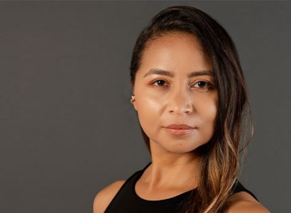 The image displays a close up of Jessica Parra, from the shoulders, up. She wears a black top. She has brown hair pulled over to the side. She looks intently into the camera. The backdrop is completely grey.