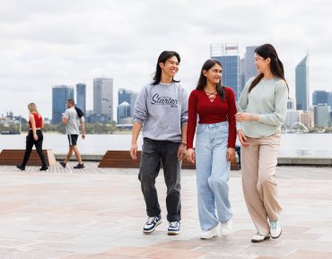 Sergio walks with two female students in South Perth. In the background is the Perth city skyline. The group of three walk towards the camera.