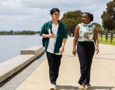 Student Cao Minh is centred in the image. He's walking alongside a female student. They are walking along a path. On one side is a body of water and on the other side is grass and trees. 