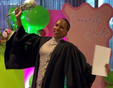 Student stands in front of the camera smiling. Her arms are lifted up, in one hand are flowers and in the other hand is her graduation certificate. She wears a graduation cloak. In the background is pink cardboard that says "Congrats Grads."