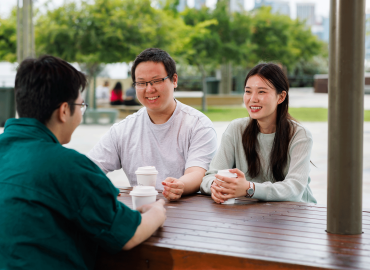 Lhaki sits at a table located in a park. She's with two other students. They all have takeaway coffees. In the background are lush green trees.