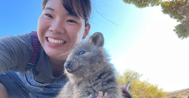 A Japanese student named Otone is taking a selfie with a friendly Quokka. The Quokka, native to Rottnest Island in Western Australia, is closest to the camera, appearing to smile. Otone is smiling at the camera as well, with the sun shining brightly in the background.