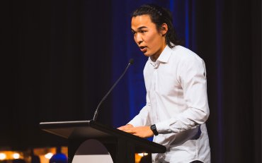 Erdene-Od, an international student from Mongolia is standing on stage looking towards an audience that is not featured in the photo. He stands on a podium, speaking into a microphone.