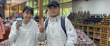 Two international students stand facing the camera, both smiling brightly. They are wearing white jackets. The young woman on the left holds a takeaway coffee cup, while the young man on the right has one hand in his jacket pocket and is making a peace sign with the other. They are inside a shop, with a display of various nougats, rocky roads, and chocolates behind them. In the distance, shelves are stocked with chutneys, sauces, and condiments.