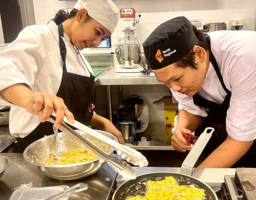 Two students stand in front of a cooking stove. On the left is student Made Della and on the right is student Minh Nguyen. Both students wear chef outfits. They are cooking together in a kitchen.