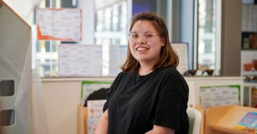 Student Athena sits on a chair facing the camera. She sits in a classroom. There's children's books sitting on a shelf and colourful posters on the wall.