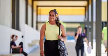 Student Keloha walking at North Metro Campus located in Perth. Keloha walks towards the camera. In the background, there's blurred students walking and sitting around the campus.