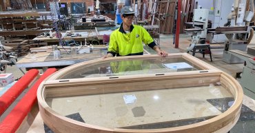 Student Pilsu Park working on a carpentry project. He wears hi-vis and a hat. He stands in front of a large circular wooden structure. He is located in a workshop with lots of tools, work benches and carpentry tools visible.