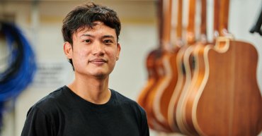 Student George (Farook). Portrait photo. He stands facing the camera. In the background, there's a line of wood guitars hanging up.