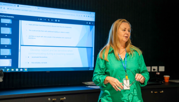 Lecturer Kylie Anne Stokes stands on the right side of the photo, looking off-camera and speaking to a group. In the background, a large PowerPoint presentation is visible.