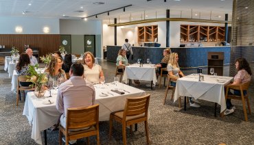 Wide shot of the training restaurant at South Metropolitan TAFE's Mandurah campus. The room is filled with neatly set tables, each covered with white tablecloths and arranged with wine glasses, cutlery, and napkins. Most tables are occupied by smiling guests. In the background, a long, sleek bar is visible, and a waiter is walking toward the kitchen.