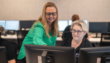  Kylie Anne Stokes stands on the left side of the photo, leaning over a student to her right and pointing at a computer screen in front of them. Both Kylie Anne and the student are smiling. In the background, more students are seated at their computer desks.