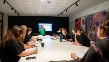 Wide shot of an Event Management class, with students seated around a long table wearing black uniforms. At the end of the table, lecturer Kylie Anne Stokes is giving a presentation.